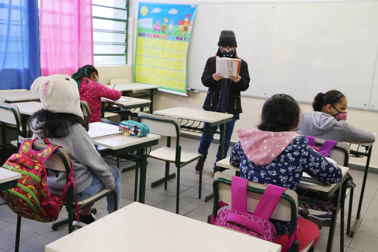 Foto mostra uma sala de aula com quatro crianças e uma professora na frente, em pé, lendo um livro.