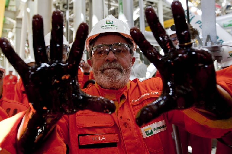 Lula aparece do peito para cima mostrando suas mãos abertas cheias de óleo. Ele veste roupas de proteção, como um capacete, óculos e macacão laranja da Petrobras. Tem barba grisalha e sorri para a câmera.