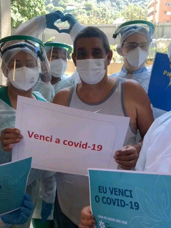 Homem branco de regata branca, usando máscara cirúrgica rodeado de enfermeiros segurando papéis com os dizeres: 'eu venci a Covid-19'