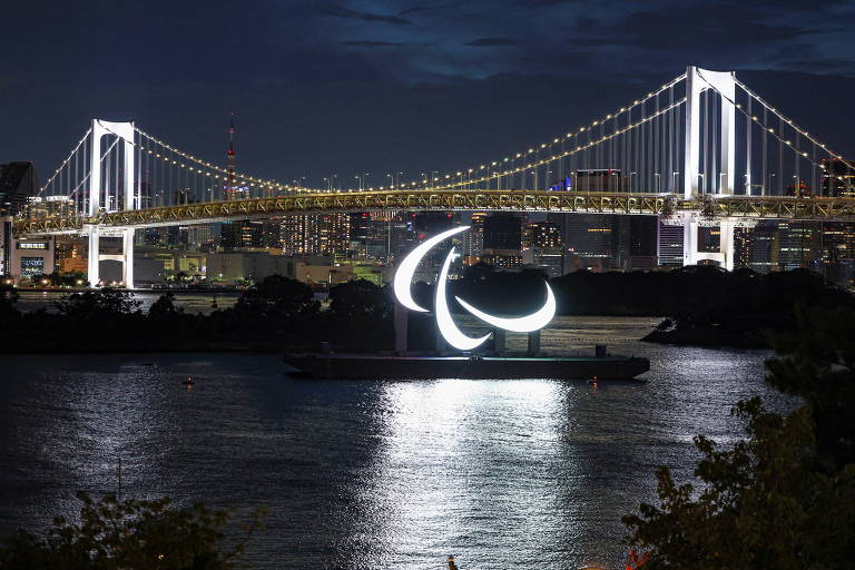 Símbolo iluminado dos Jogos Paralímpicos de Tóquio-2020 em uma baia, com uma ponte ao fundo, durante a noite