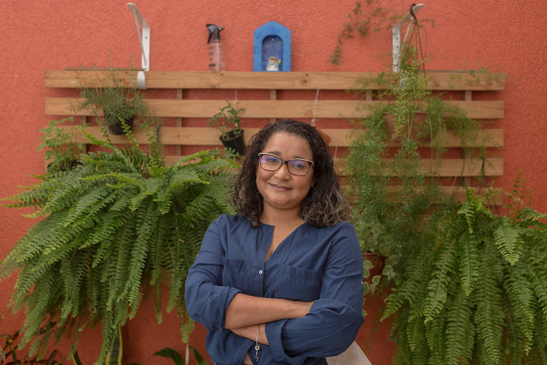 Mulher vestindo camisa azul com os braços cruzados em frente a um estrado de cama adaptado para um jardim vertical, pendurado em uma parede salmão