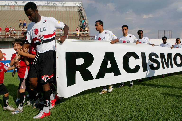 Jogadores do São Paulo, liderados pelo atacante Grafite, entram em campo com faixa contra o racismo. Grafite está com cabeça abaixada e com o uniforme branco do São Paulo. A faixa que ele segura à frente é branca e tem o dizer "Racismo" em preto