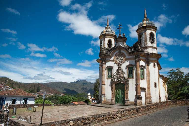 A imagem captura a fachada de uma igreja barroca, com suas duas torres ornamentadas e simétricas sob um céu azul claro. A arquitetura detalhada contrasta com a simplicidade das montanhas ao fundo, enquanto uma praça de pedras completa o ambiente