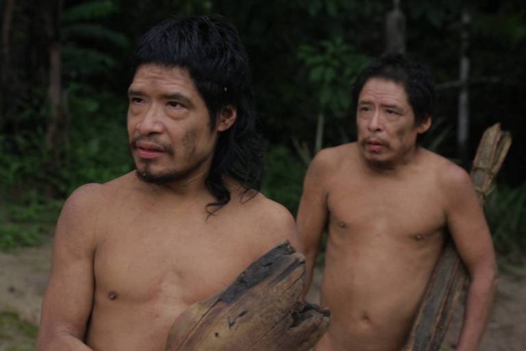 Dois homens indígenas sem camisa estão de pé diante de árvores grandes