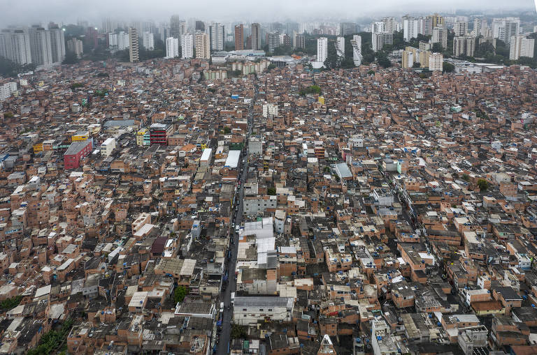 Foto mostra vista área da favela de Paraisópolis. No fundo, é possível ver vários prédios e árvores. O céu está nublado e com neblina.
