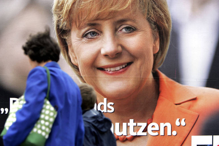 Mulher carregando uma sacola sob o braço esquerdo e levando uma criança pela mão direita passa em frente a cartaz com foto de Merkel, de paletó laranja e sorrindo