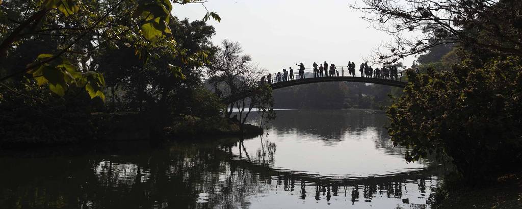 Vemos um lago, com árvores dos dois lados; ao fundo, sobre ele, uma ponte em arco, cheia de gente, cujo reflexo se vê na água