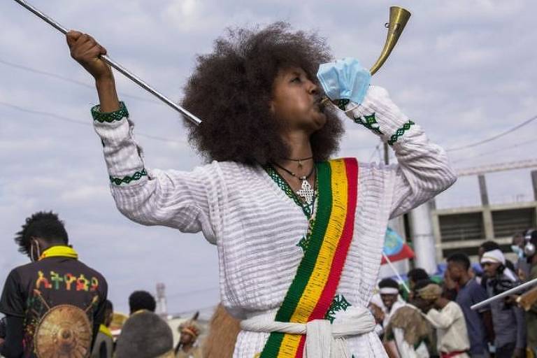 mulher toca instrumenyo de sopro e tem uma faixa com as cores verde, amarela e vermelha da bandeira etíope