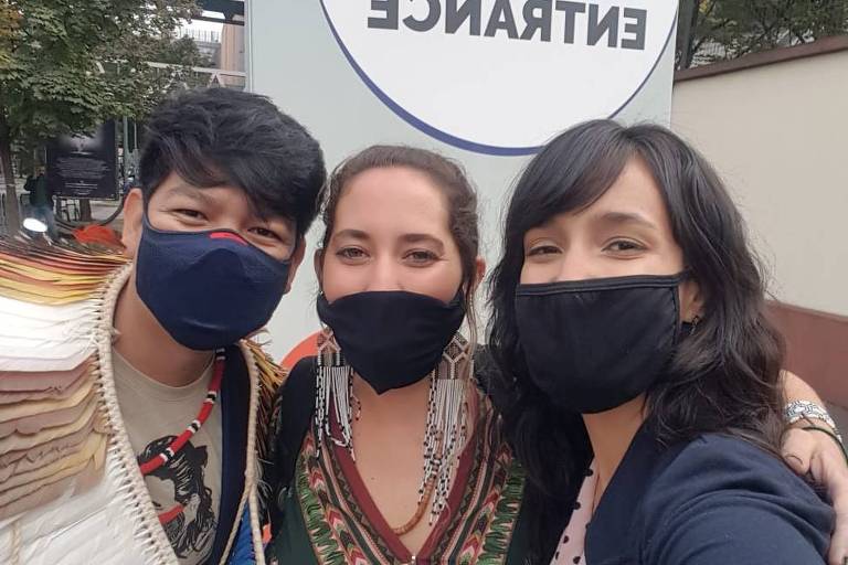 Três jovens se abraçam em frente a cartaz