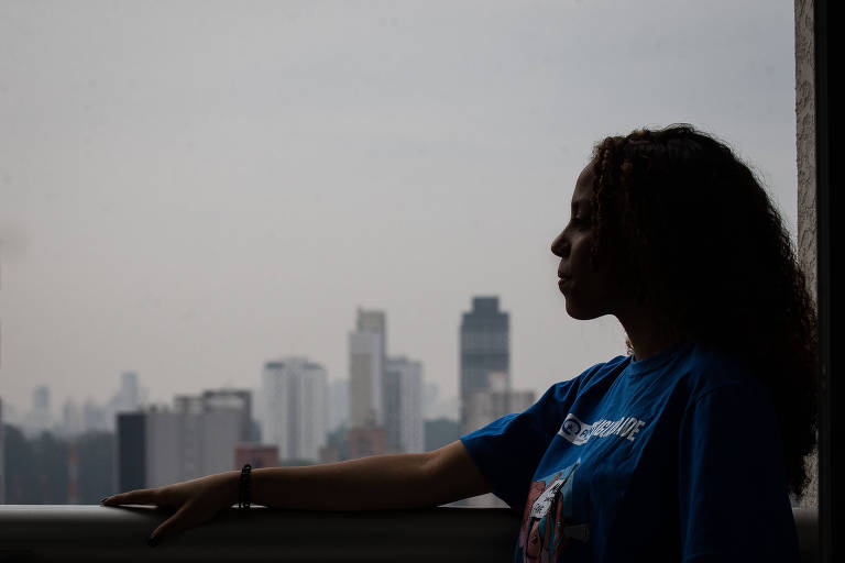 Silhueta de uma mulher negra de perfil, com camiseta azul, no canto direito da janela com a vista de São Paulo ao fundo. O céu está nublado e existem poucos prédios na parte inferior da foto, logo acima do batente da janela