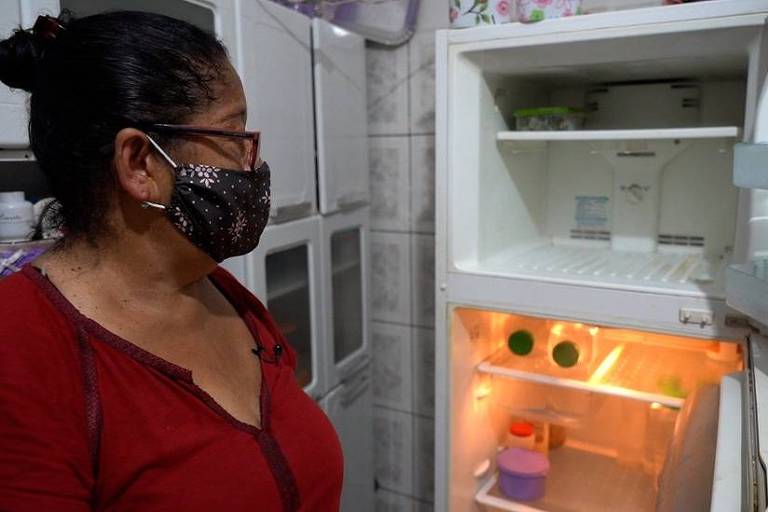 Mulher negra, de óculos, máscara escura e camisa vinho olha para sua geladeira branca e vazia