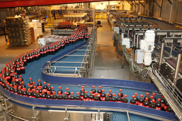 Imagem mostra interior de uma fábrica de refrigerantes. Entre os maquinários, se destaca uma esteira azul em formato de "S", cheia de garrafas de Coca-Cola.