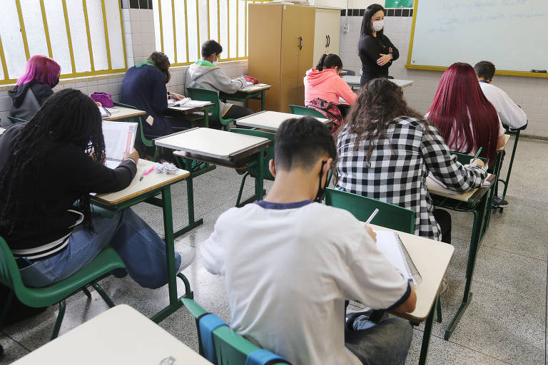 Alunos sentados em carteiras dentro de uma sala de aula, com uma professora ao fundo