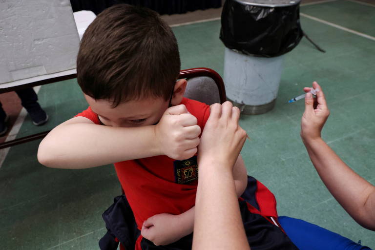 Criança tapa os olhos com o braço enquanto um profissional de saúde aplica a vacina contra Covid