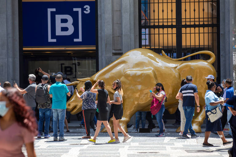 Pessoas se aglomeram ao redor de uma escultura dourada em formato de touro em frente à fachada da Bolsa de Valores de São Paulo, onde é possível ver um [B]³ escrito.