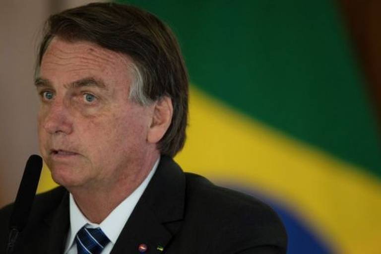 Imagem em primeiro plano mostra Bolsonaro de social. Ao fundo, se vê uma bandeira do Brasil