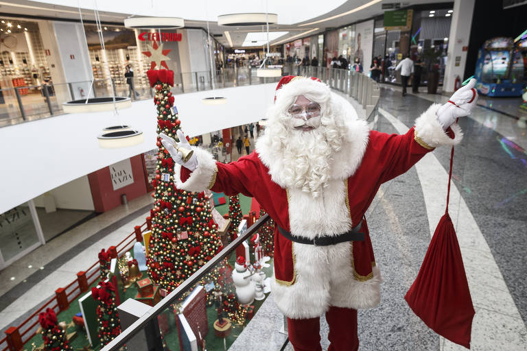 Fotografia colorida do Papai Noel em pé, de braços abertos e segurando um saco vermelho na sua mão esquerda. No fundo, há uma árvore de natal.