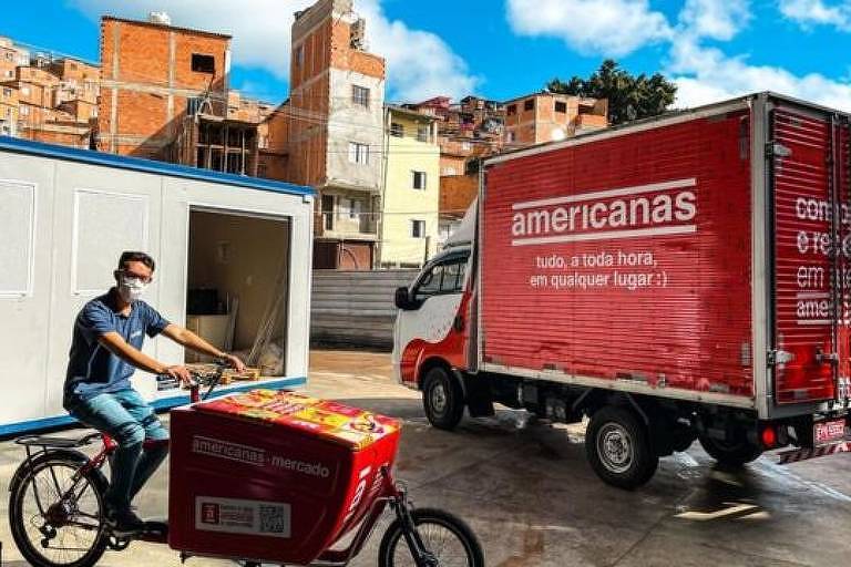 Um homem de máscara está em cima de uma bicicleta. Ao fundo se vê um caminhão das Americanas e casas