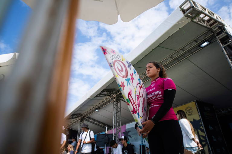 Sophia, com lycra rosa, segura a prancha debaixo do braço e tem a expressão concentrada