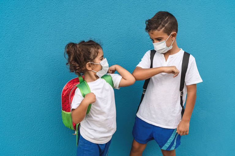 Fotografia colorida de duas crianças em pé em frente a uma parede azul. Eles usam uniforme escolar e se cumprimentam, encostando os cotovelos. Do lado esquerdo, a menina aparenta ter 3 anos e é mais baixa que o menino da esquerda. Ela usa mochila com desenho de melancia e ele usa mochila com alças escuras. Os dois tê pele morena, cabelos castanhos escuros estão de máscara de proteção.  