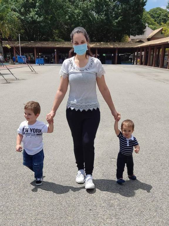 Claudia é branca, está em pé, com cabelo amarrado, vestindo blusa clara e calça escura. Está entre os filhos Heitor, de dois anos e Davi, de um. Ela usa máscara de proteção.