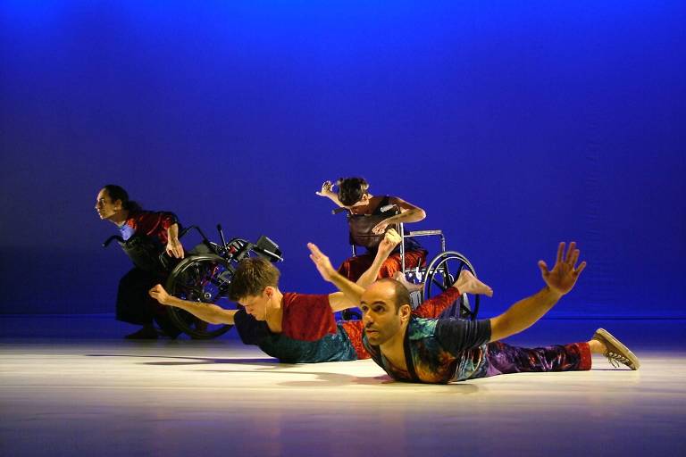 Performance de palco mostra quatro pessoas, duas em primeiro plano deitadas no chão com os braços estendidos e duas ao fundo, em cadeiras de roda, inclinando o corpo para frente