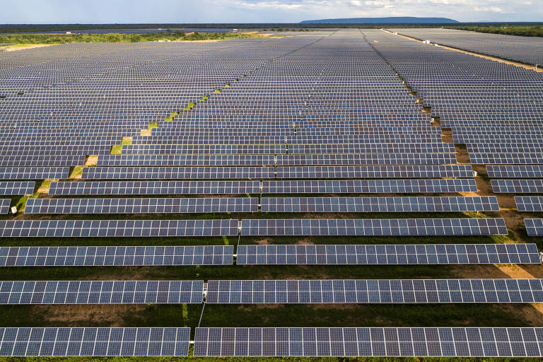 A imagem mostra um vasto campo de painéis solares dispostos em fileiras organizadas. O céu está parcialmente nublado e a paisagem ao fundo é plana, com vegetação rasteira. Os painéis solares ocupam a maior parte da imagem, indicando uma grande instalação de energia solar.