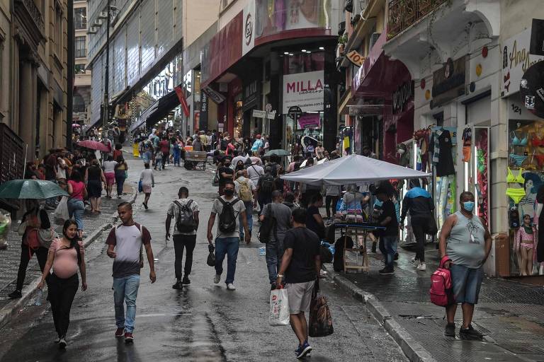 Pessoas andando em ladeira repleta de lojas, durante chuva, algumas com guarda-chuva