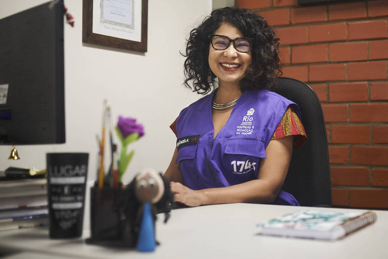 Mulher de cabelos encaracolados, vestindo um colete roxo, está sentada em frente a um computador e sorri, olhando para frente