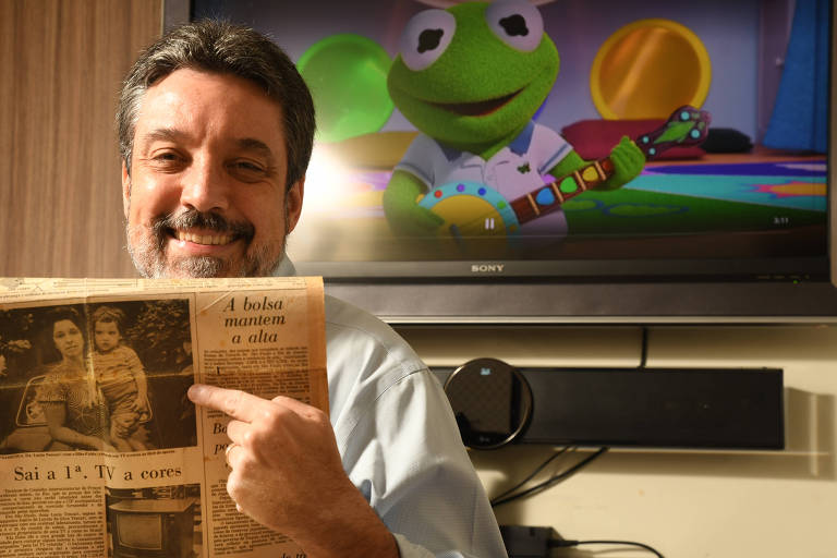 Homem branco, de barba, tendo TV ligada ao fundo, com a transmissão de um programa infantil, segura um jornal antigo
