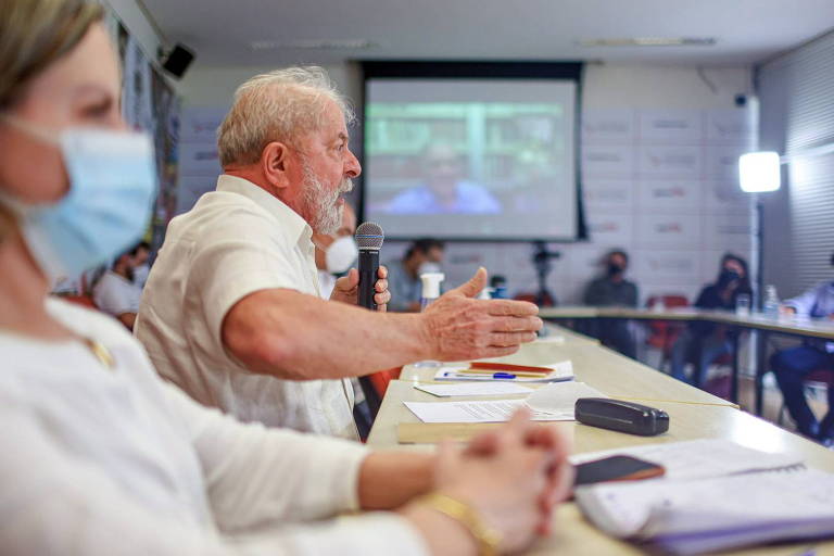 Homem de barba e cabelos brancos, com camisa clara, gesticulando ao falar numa reunião. Ao seu lado, uma mulher jovem, loira, de blusa branca e máscara de proteção contra o coronavírus. Veem-se outros participantes da reunião ao fundo e num telão instalado para os que participam remotamente. 