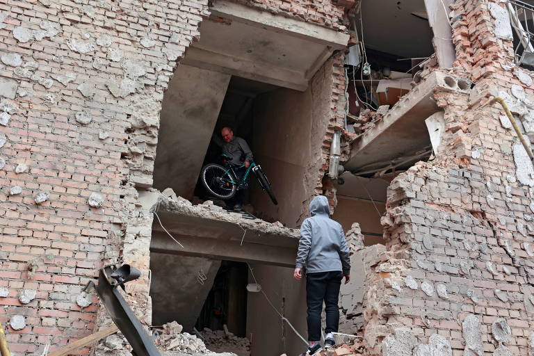 De costas, vestida de calça azul escura e blusa azul clara, mulher observa homem que carrega uma bicicleta dentro de um prédio de tijolos com as paredes destruídas