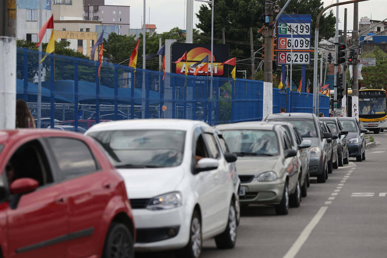 Fila de carros de diversas cores para abastecer em posto de combustíveis. Há um carro vermelho na frente, um branco atrás e vários prateados