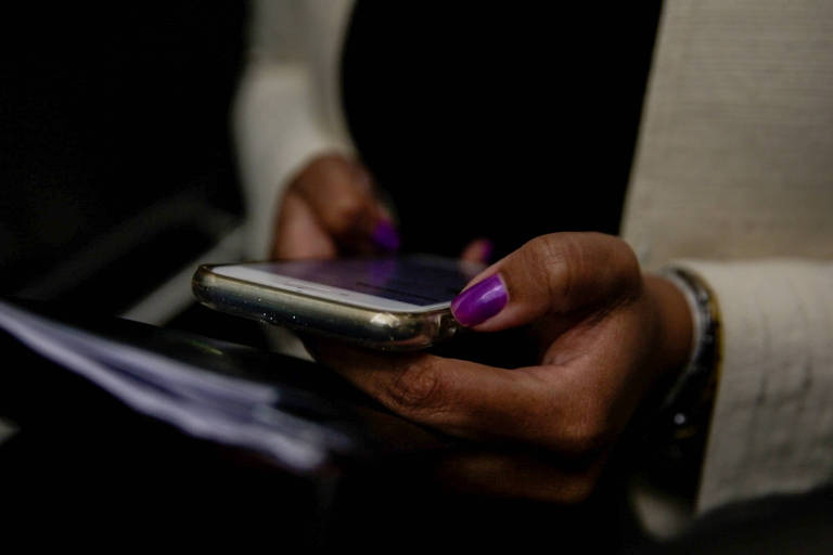 Mulher negra, com as unhas pintadas de roxo, mexe em celular prata