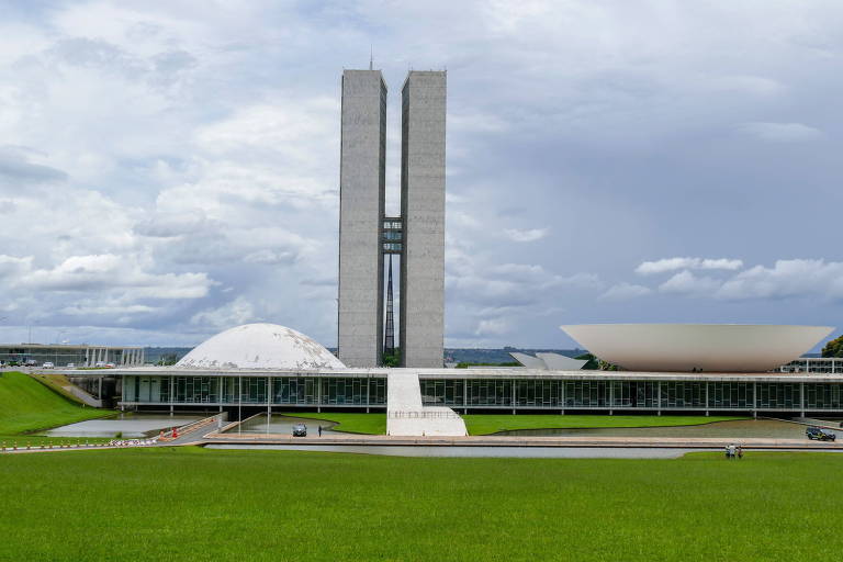Imagem mostra sede das duas Casas do Poder Legislativo brasileiras. Há duas cúpulas, uma côncava e outra convexa, além de duas torres com 100 metros. Os prédios são cinza e estão em frente a um gramado verde.