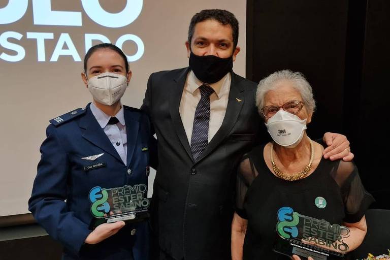 Imagem colorida mostra três pessoas, da cintura para cima; à esquerda, mulher branca, de uniforme azul e máscara branca, segurando um troféu de acrílico; no centro, homem branco, de terno escuro, gravata e camisa branca; à direita, mulher branca, idosa, de cabelos brancos e máscara branca, segurando um troféu de acrílico.