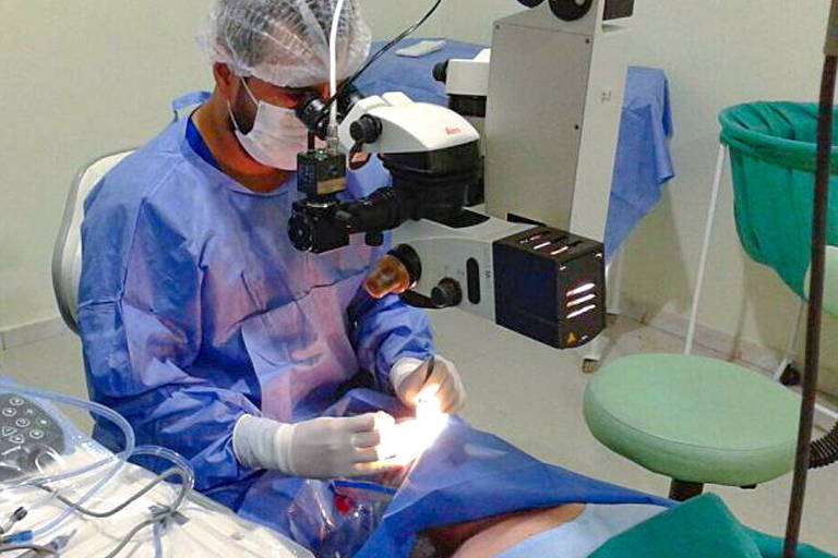 detalhe de sala cirúrgica, com médico de avental e touca operando paciente em maca