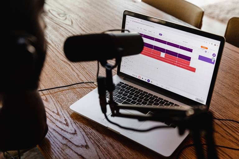 Foto mostra laptop em cima de uma mesa de madeira. A tela é branca, com algumas linhas em roxo e vermelho. Em primeiro plano, está uma mulher, de costas, com um fone de ouvido. Na frente dela, há um microfone, sustentado por um suporte.