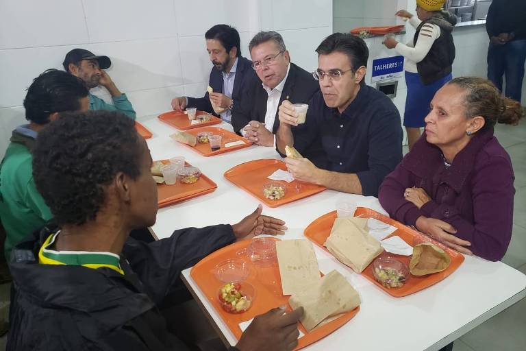 À mesa, sete pessoas, só uma mulher, com bandejas laranjas, com salada de fruta, café e um pequeno pacote marrom, diante de si. Rodrigo Garcia, um homem branco, cabelos castanhos, óculos e camisa azul escura, segura o pão em uma mão e um copo de plástico com café com leite na outra. Ele está ao lado de uma mulher com blusa roxa, que observa um rapaz negro de blusa preta que está sentado diante dela falando com Garcia, que olha pra ele enquanto mastiga. Do lado de Garcia, Carlão, um homem branco, cabelos grisalhos e blusa preta, também observa. Do lado dele, Ricardo Nunes, um homem branco, de barba e cabelos escuros, com blusa cinza, está comendo e olhando para a bandeja. Diante dele, um rapaz negro de boné tem uma das mãos apoiada no rosto e olha em direção ao prefeito