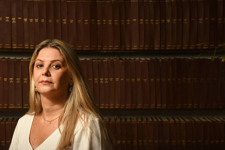 Mulher loira, de camisa branca, tendo livros de uma biblioteca ao fundo