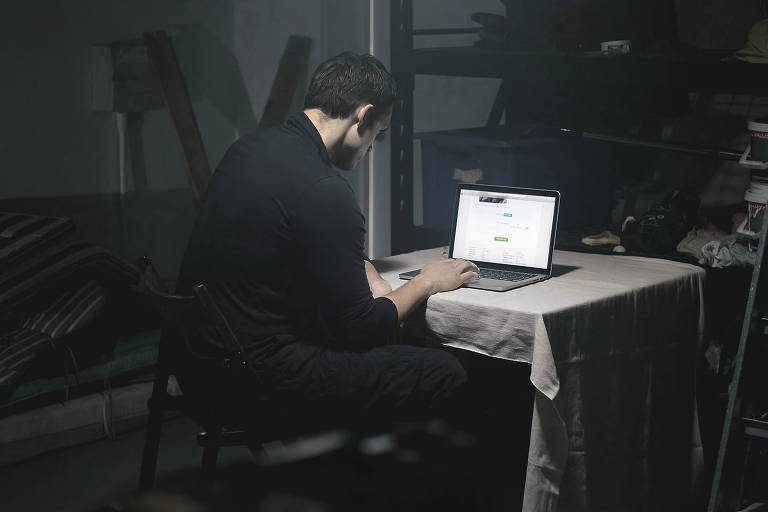 Um indivíduo trabalha em um laptop em um ambiente escuro e isolado, iluminado apenas pela tela do computador