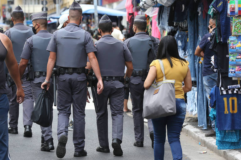 Imagem colorida mostra cinco policiais militares de costas, de corpo inteiro, andando juntos em uma rua. Eles vestem um uniforme na cor cinza, e todos usam um quepe cinza.