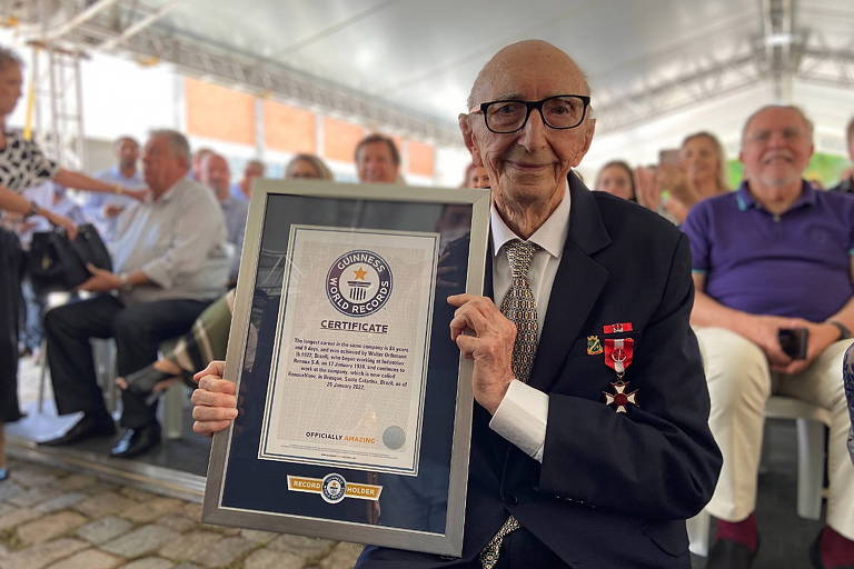 homem idoso com terno e gravata segura placa do Guiness Book com mensagem em inglês de funcionário mais antigo do mundo, com pessoas atrás em um evento