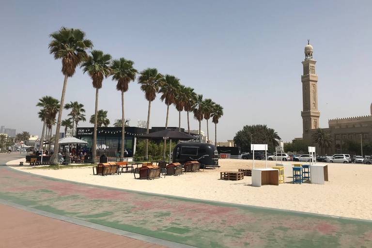 Mesas e cadeiras na areia da praia, ao lado de palmeiras, com uma mesquita ao fundo