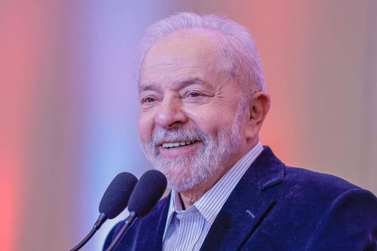 Em foto colorida, Lula aparece em plano médio vestindo terno azul escuro e camisa clara; ele tem as mãos entrelaçadas e fala sorrindo diante de um microfone
