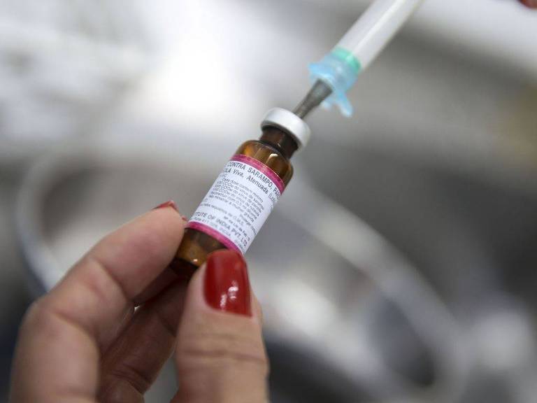 Mão com unhas pintadas de vermelho segura frasco de vacina contra o sarampo enquanto uma seringa extrai o líquido do recipiente