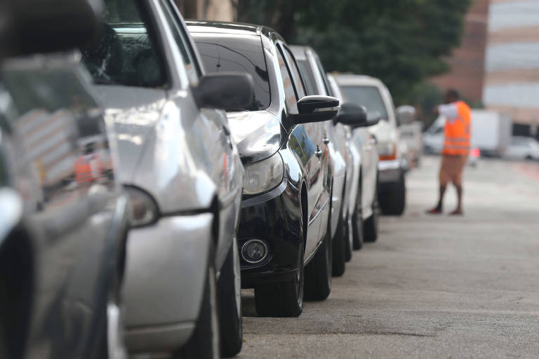 Fileira de carros estacionados à esquerda com homem de colete laranja ao fundo