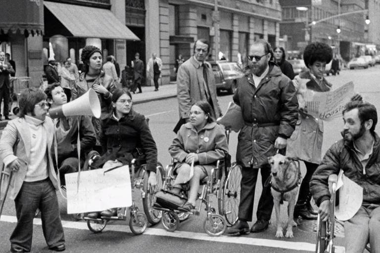 Em uma imagem em preto e branco, várias pessoas com deficiência fazem protesto em uma rua. Uma delas, com uma muleta, usa um megafone. Há cadeirantes com cartazes nas mãos e uma pessoa com um cão-guia