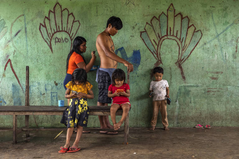 Grupo com dois adultos e três crianças faz pinturas na pele diante de uma parede verde com desenhos de cocar