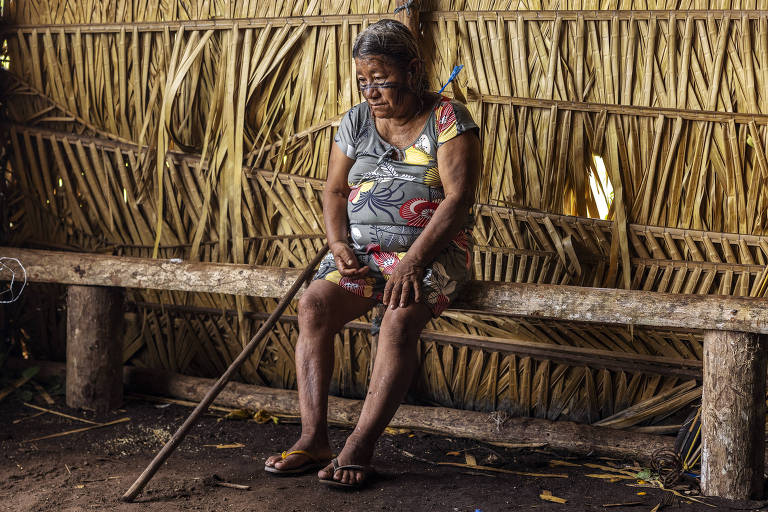 Mulher idosa com vestido colorido e rosto pintado com tinta preta está sentada em um banco de madeira dentro de uma casa feita de palha